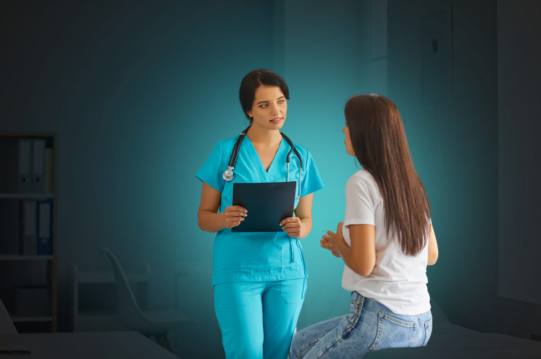 A healthcare professional in blue scrubs holding a clipboard and having a conversation with a patient seated on an examination table in a medical setting.